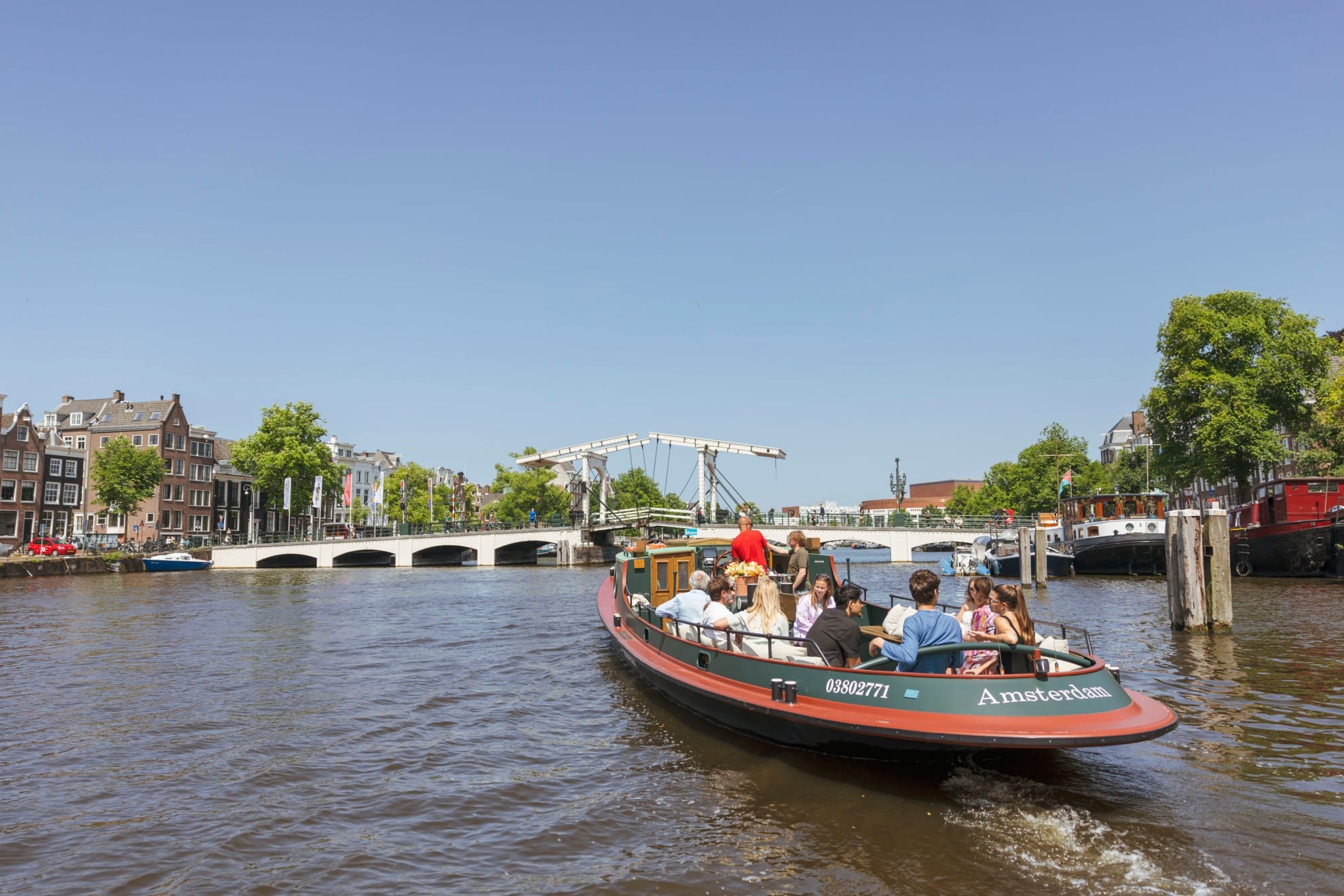 Un barco clásico lleno de pasajeros navega por un canal de Ámsterdam hacia el Magere Brug en un día soleado.